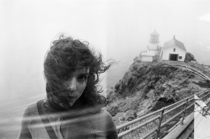 A woman with windswept hair is in the foreground of a hazy landscape with a lighthouse on a cliff.