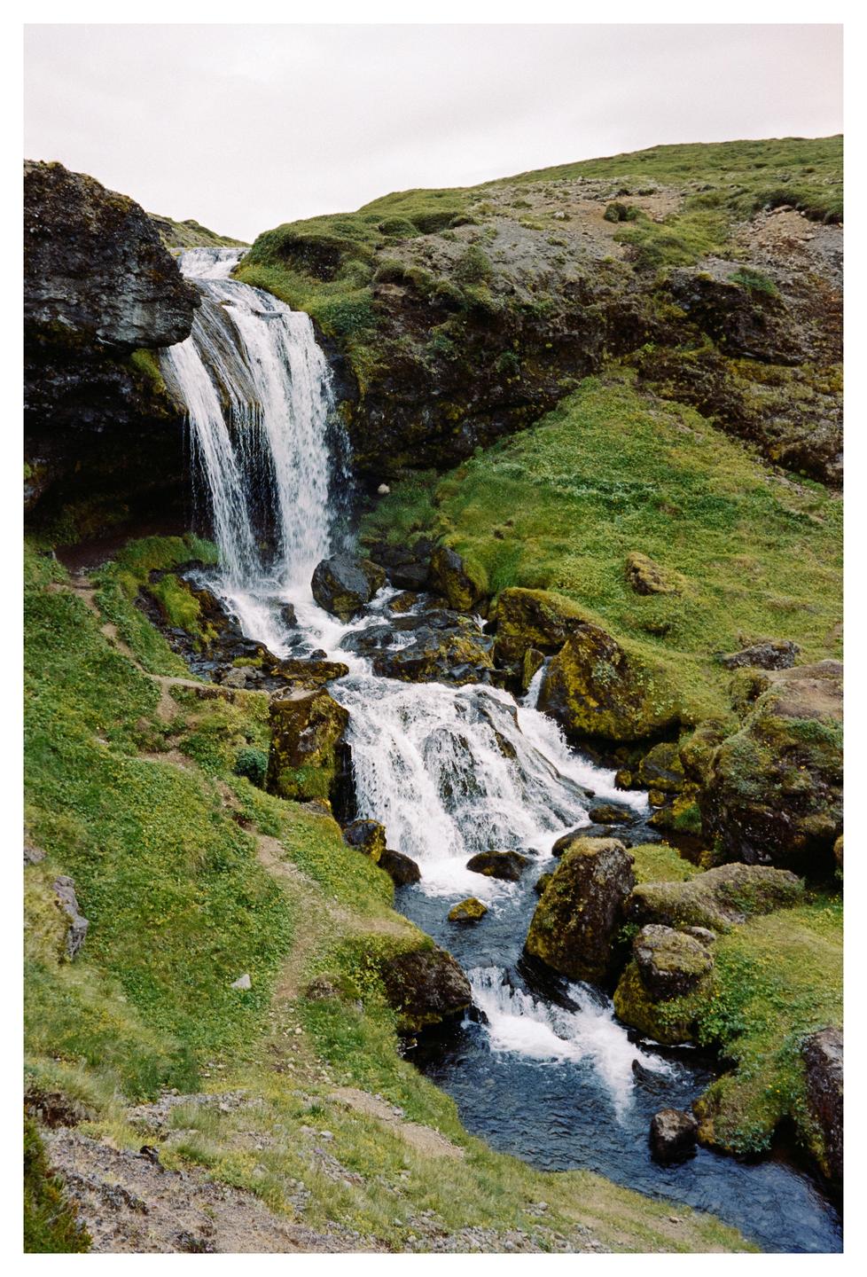 A waterfall cascades down a mossy, rocky hillside with a sheep visible near the top of the falls.