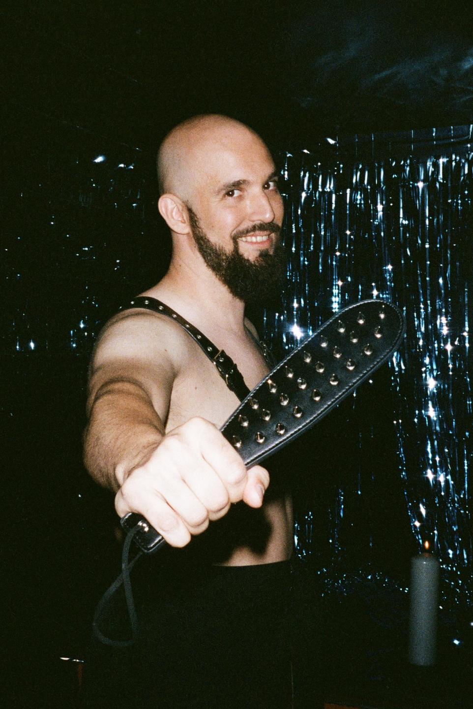A man with a beard and bald head smiles at the camera while holding a studded paddle.