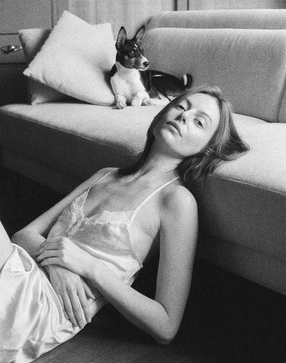 A woman in a slip lies on the floor with a dog on the couch behind her.