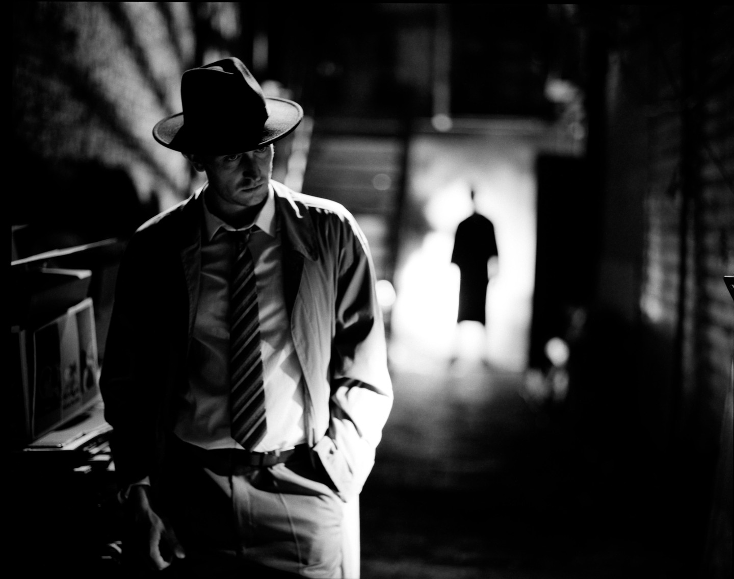 A man in a fedora and trench coat stands in a dark alleyway with another figure in the background.