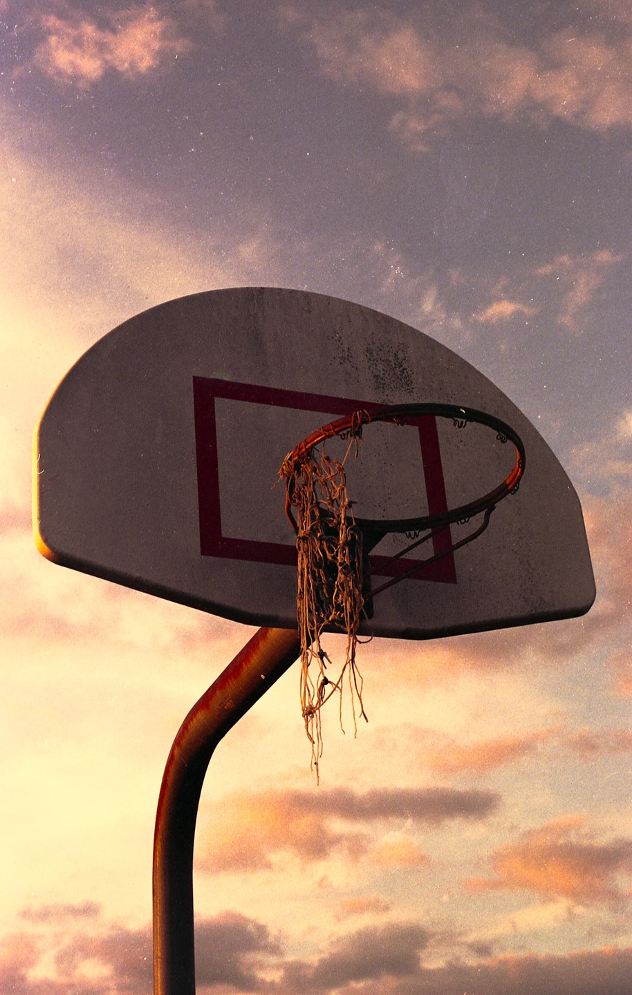 A basketball hoop with a tattered net is silhouetted against a cloudy sky at dusk.