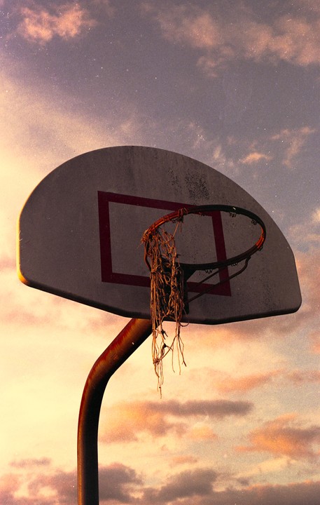 A basketball hoop with a tattered net is silhouetted against a cloudy sky at dusk.
