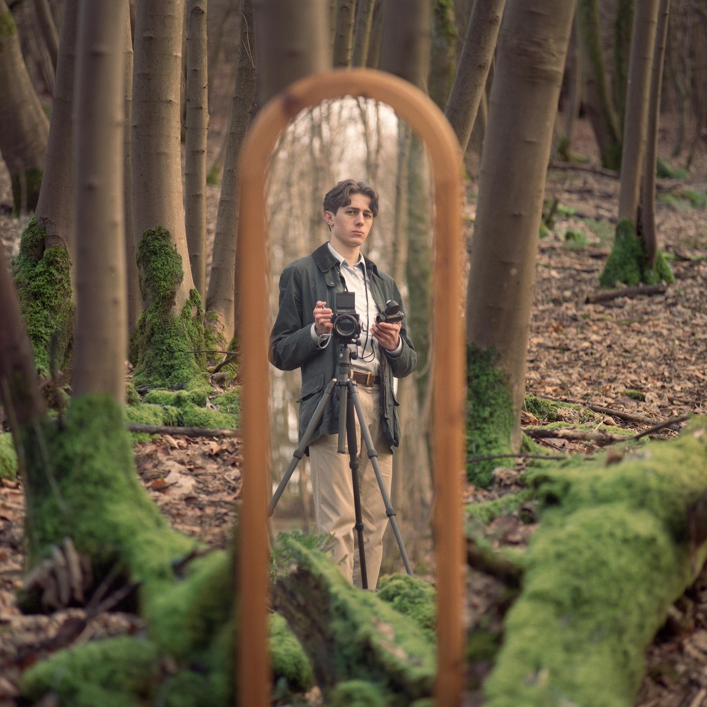 A man holding a camera on a tripod is reflected in a mirror in a forest.