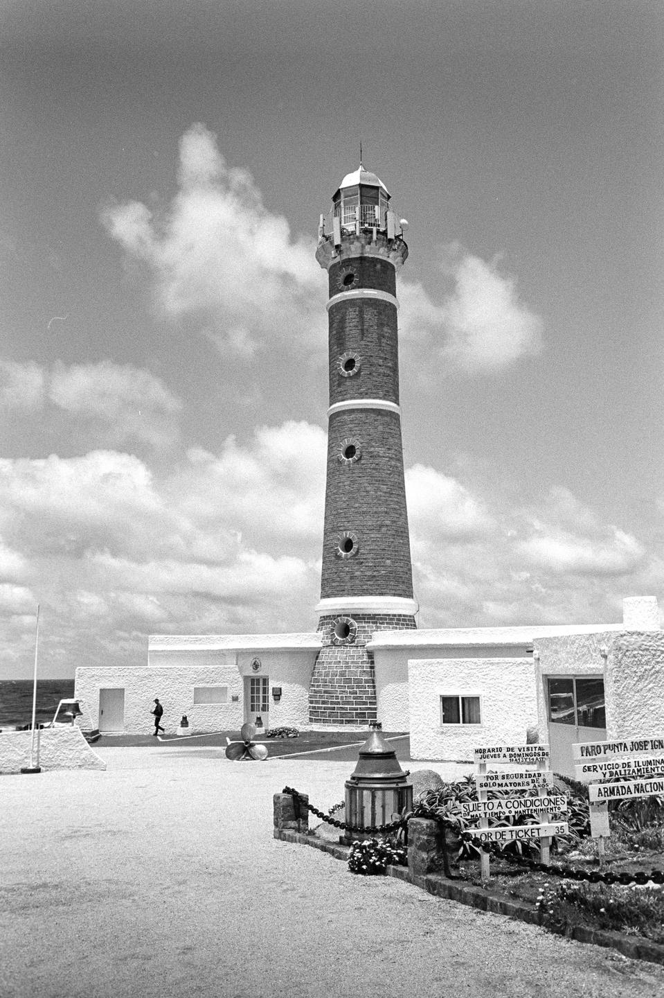 A tall lighthouse stands next to a building with a person walking nearby, all under a cloudy sky.