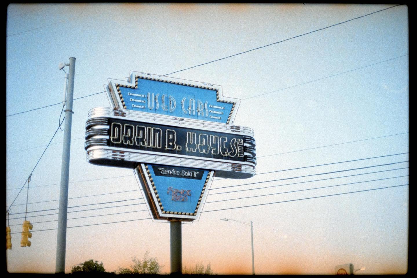 A blue and silver sign for "Used Cars" and "Orrin B. Hayes" stands against a clear sky.