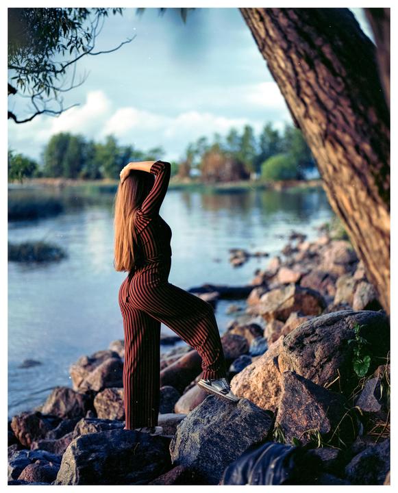 A woman in a striped jumpsuit stands on rocks by a lake, with her back to the viewer.
