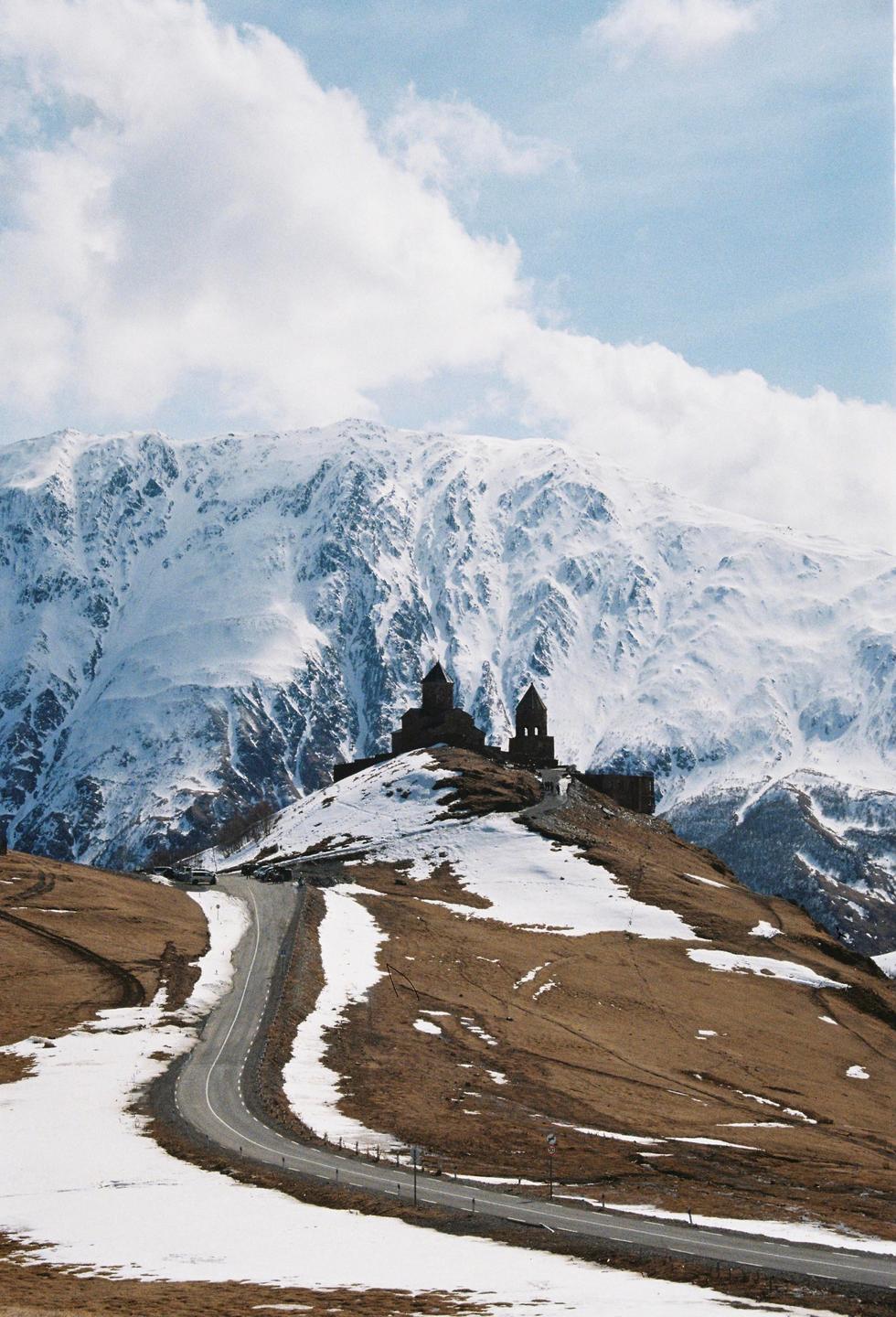 A winding road leads up a snowy hill to a church with a large snow-covered mountain in the background.