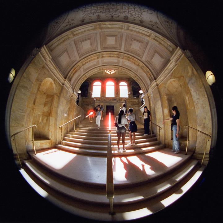A group of people stand on a grand staircase with a glowing red light in the center and three arched windows at the top.