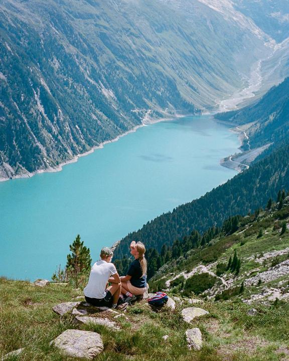 A couple sits on a grassy mountain overlooking a turquoise lake surrounded by mountains.