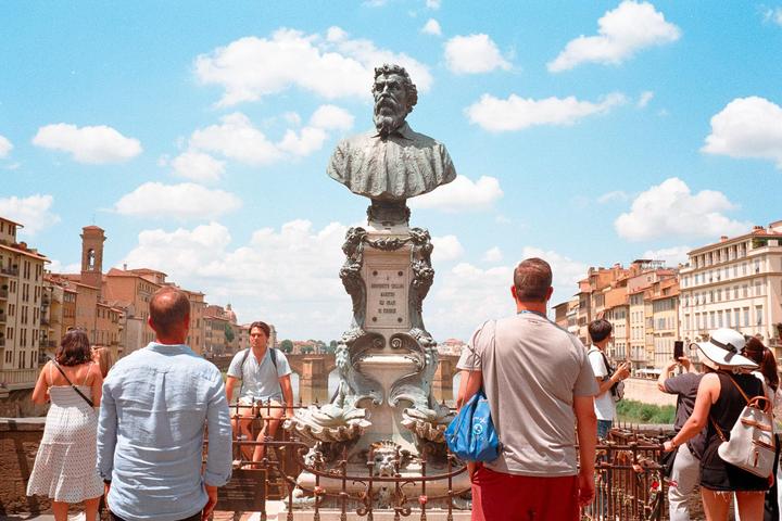 A bust of a man on a pedestal stands on a bridge with people looking at it and the buildings in the background.