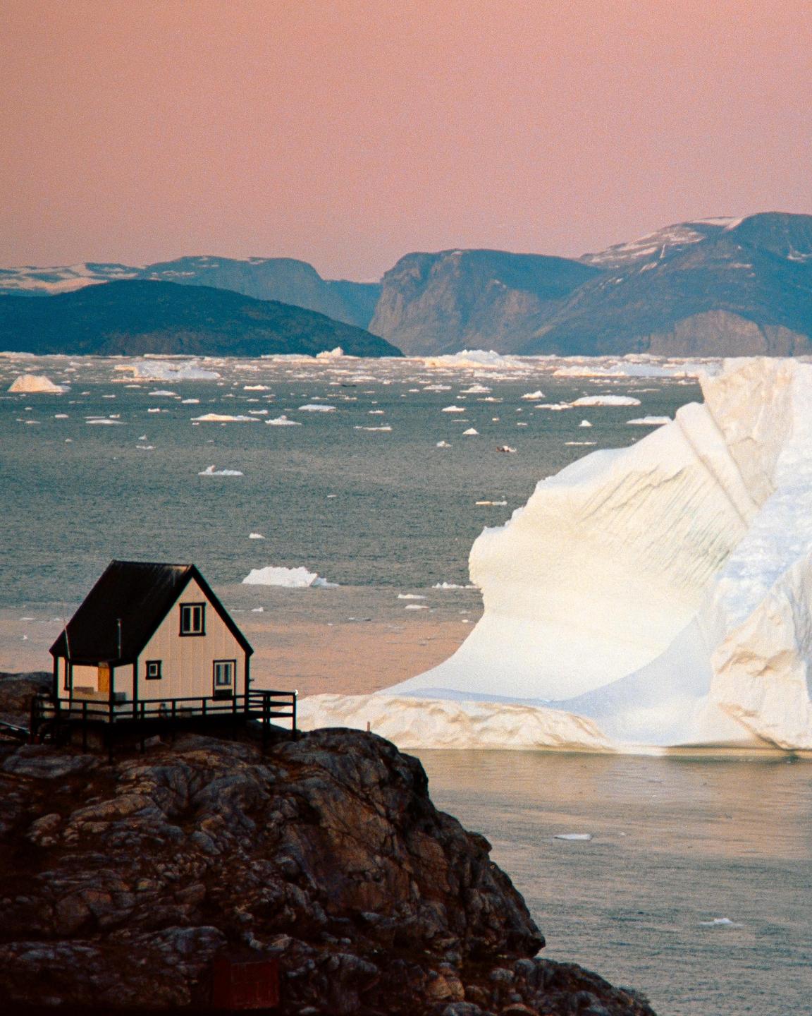 A small house sits on a rocky cliff overlooking a body of water filled with icebergs and mountains in the distance under a pink sky.