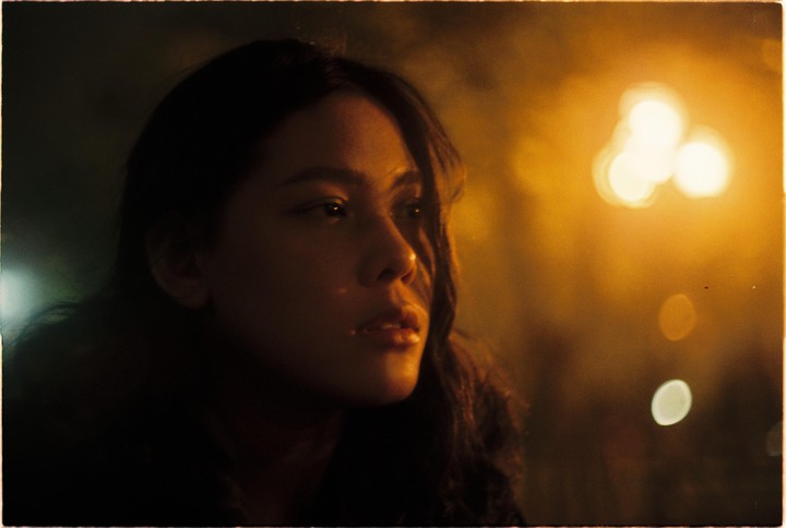 A woman's face is illuminated by a warm glow from the right, with bokeh lights in the background.