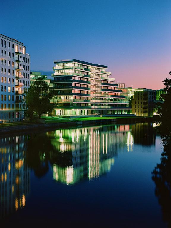 Modern buildings with illuminated windows are reflected in the water at dusk.