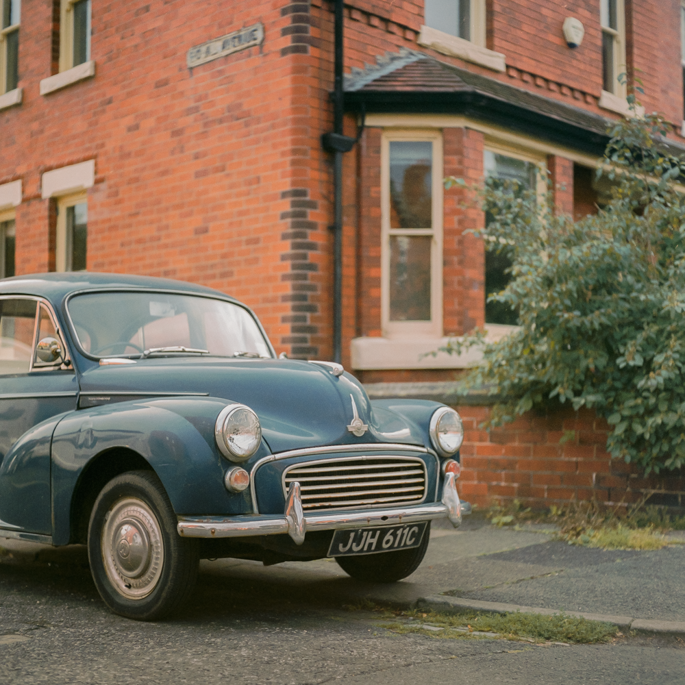 A blue vintage car is parked on a residential street in front of a brick house with a bay window.