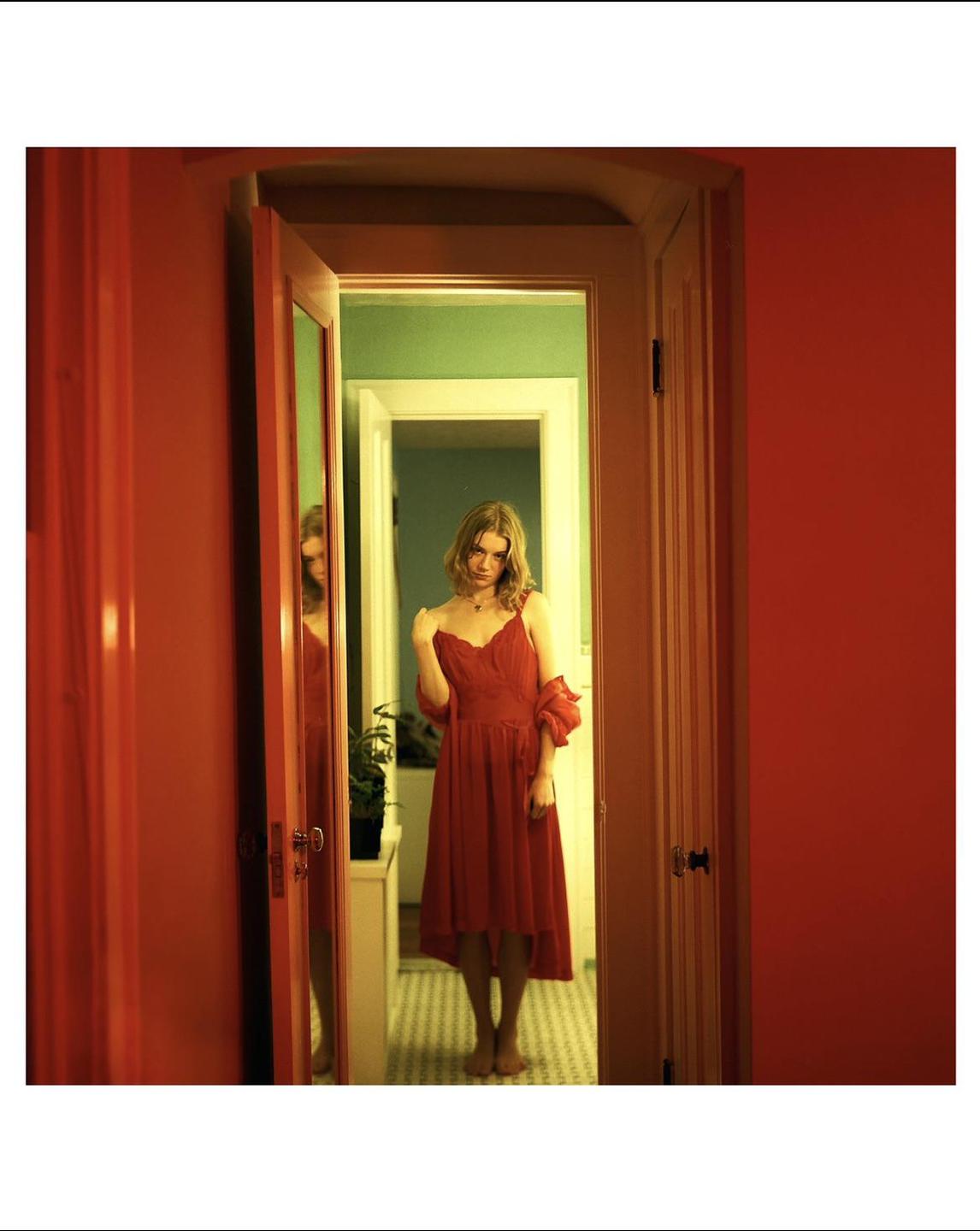 A woman in a red dress stands barefoot in a doorway, with a reflection visible in a mirror to her left.
