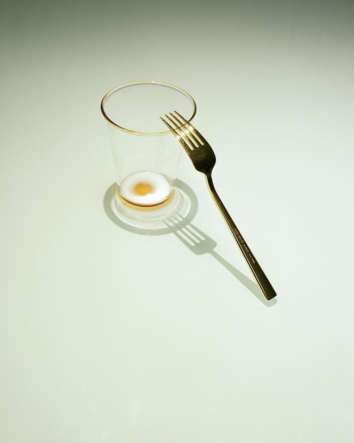 A golden fork rests on the rim of a glass containing a small amount of liquid with a yellow center.