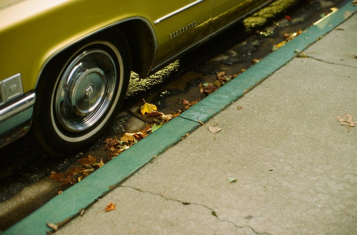 A yellow car is parked next to a green curb with autumn leaves scattered on the ground.