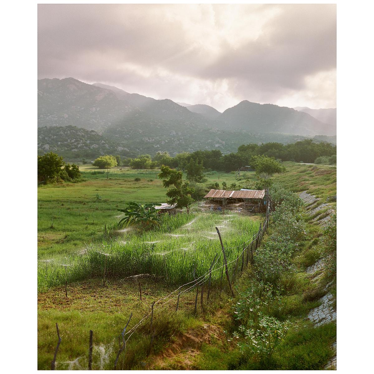 A small shack with a rusty roof sits in a green field with mountains in the background under a cloudy sky.