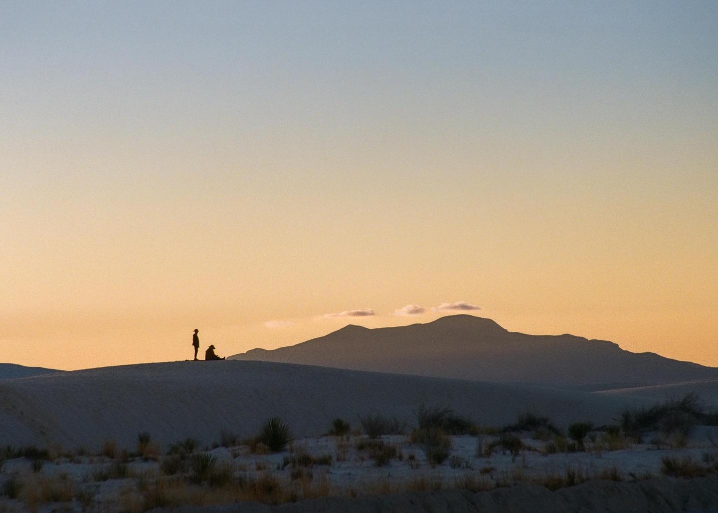 Two people stand on a sand dune with mountains in the background at sunset.