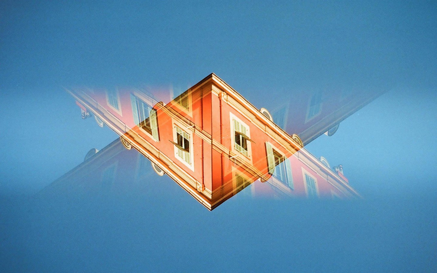 A double exposure shows a red building with white window frames and green shutters against a blue sky, with a mirrored, faded image of the building overlapping it.