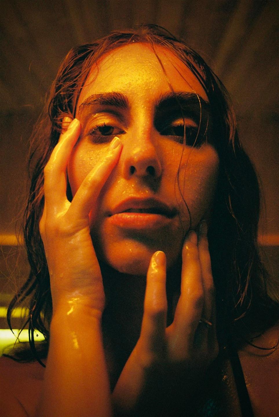 A woman with wet hair and skin looks directly at the viewer, with her hands touching her face.