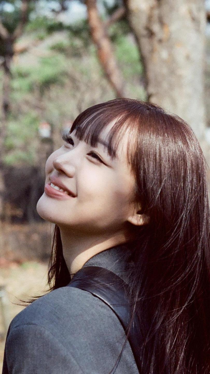 A woman with long hair and bangs smiles while looking up, with trees in the background.