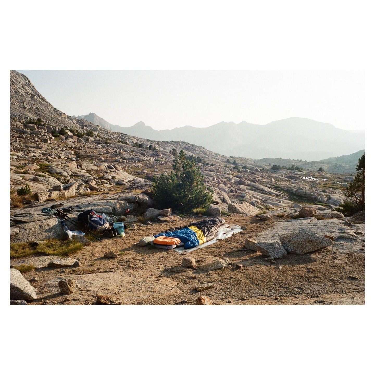 A sleeping bag and backpack are laid out on a rocky, mountainous landscape with a hazy sky.
