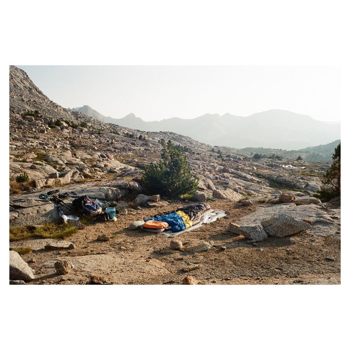 A sleeping bag and backpack are laid out on a rocky, mountainous landscape with a hazy sky.