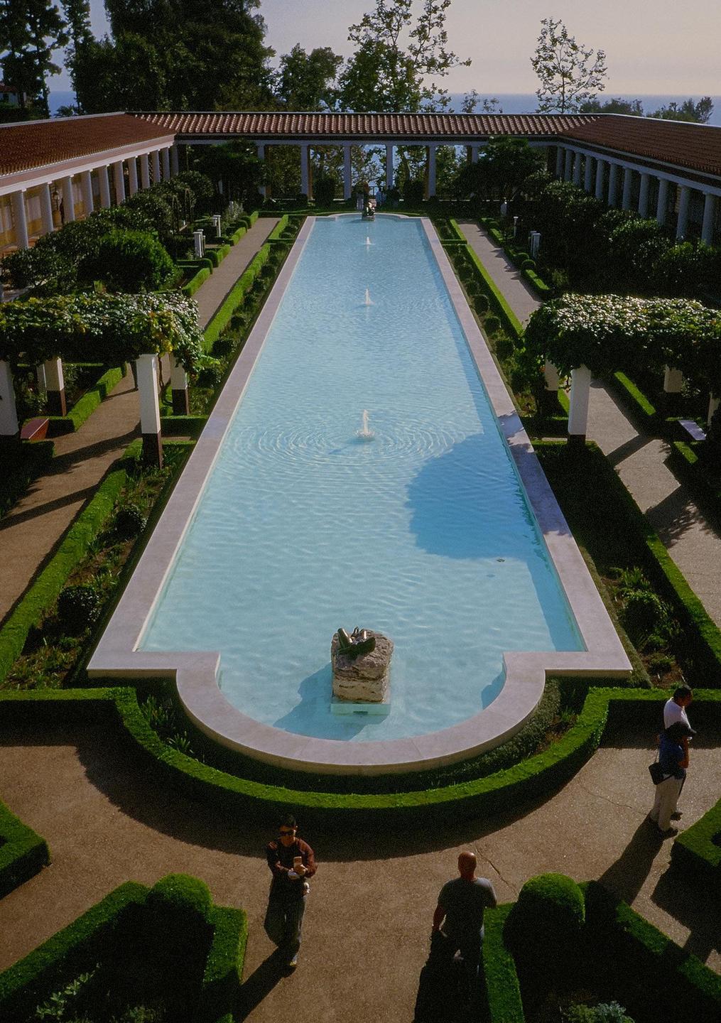 A long, rectangular pool with fountains is surrounded by manicured gardens and classical architecture under a clear sky.