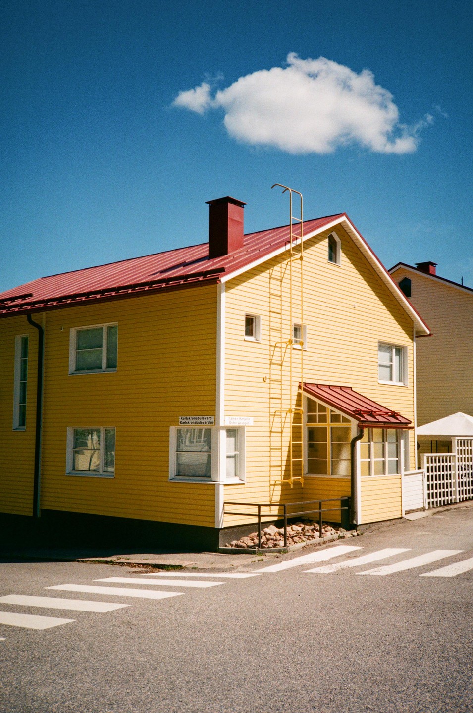 A bright yellow house with a red roof stands at a street corner under a blue sky with a single cloud.