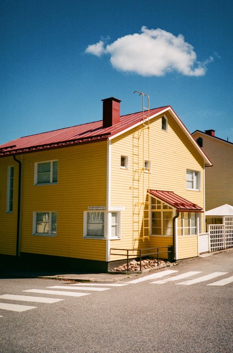 A bright yellow house with a red roof stands at a street corner under a blue sky with a single cloud.