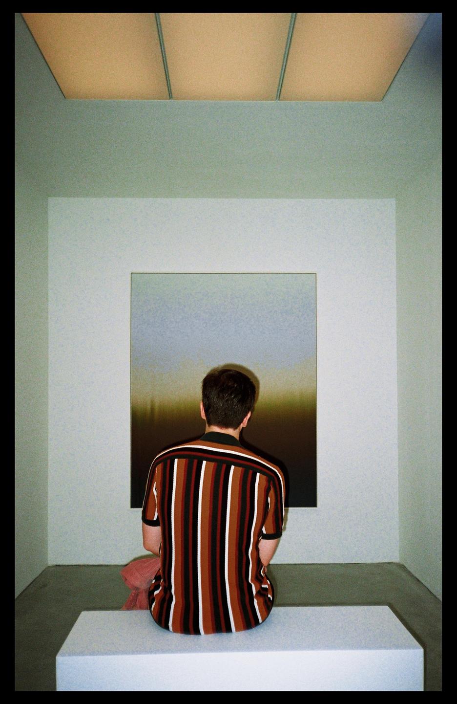 A man sits on a bench facing a painting in a museum.