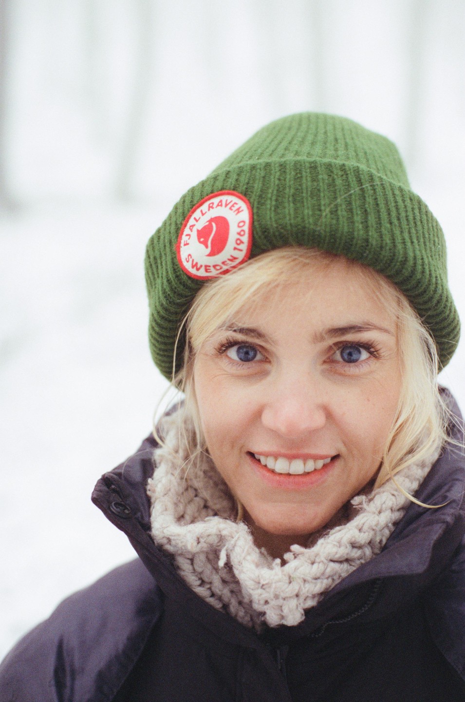 A woman wearing a green beanie and a knitted scarf smiles at the viewer in a snowy setting.