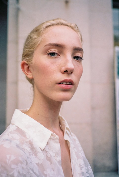 A young woman with blonde hair and a sheer white floral shirt looks directly at the viewer.