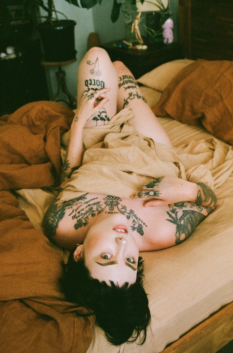 A woman with many tattoos is lying on a bed with her head at the bottom of the frame, looking up at the viewer.