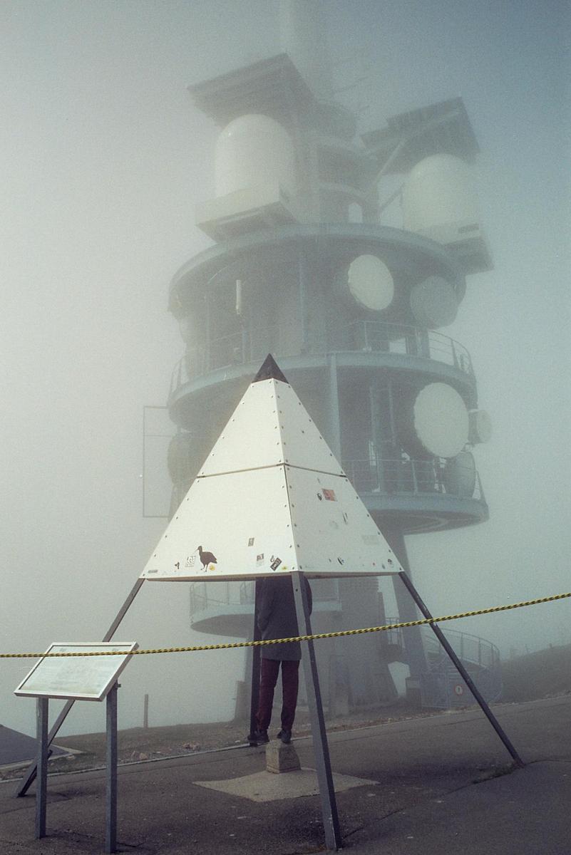 A person stands on a concrete block under a white pyramid structure, with a large building shrouded in fog behind them.