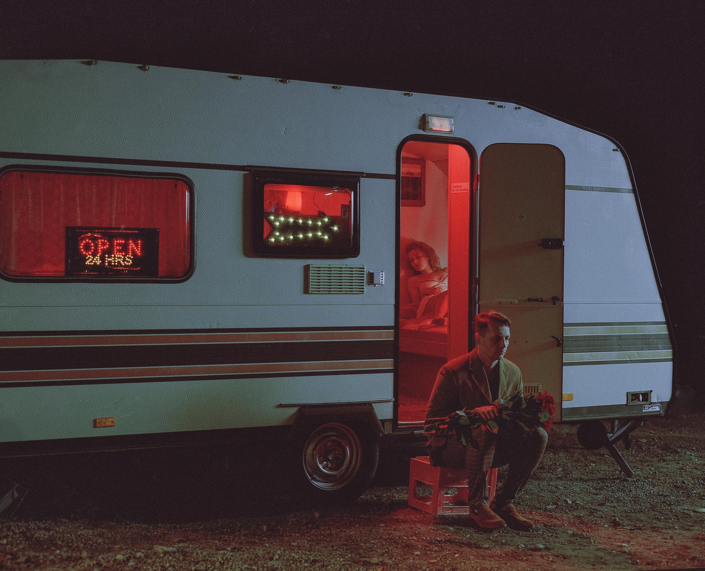 A man sits outside a trailer with a bouquet of roses while a woman is visible inside through the open door, illuminated by red light.
