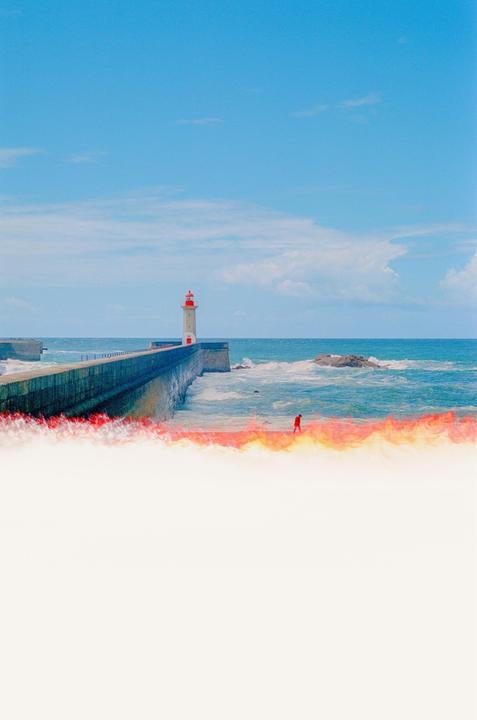 A lighthouse stands at the end of a long pier with a person walking along the water's edge.