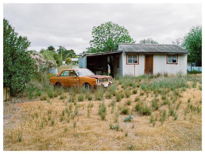 An abandoned orange car sits in a grassy field next to a small white building under a cloudy sky.