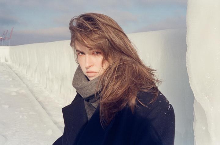 A woman with long hair and a scarf looks at the viewer, standing next to an ice-covered wall.