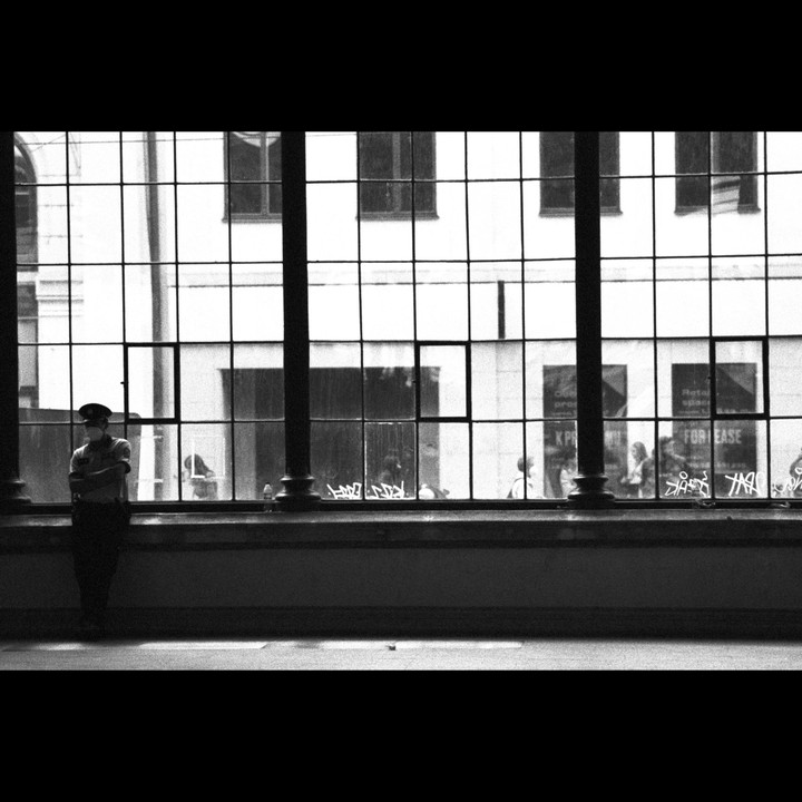 A man in uniform stands in front of a large window with many panes, with people and buildings visible outside.