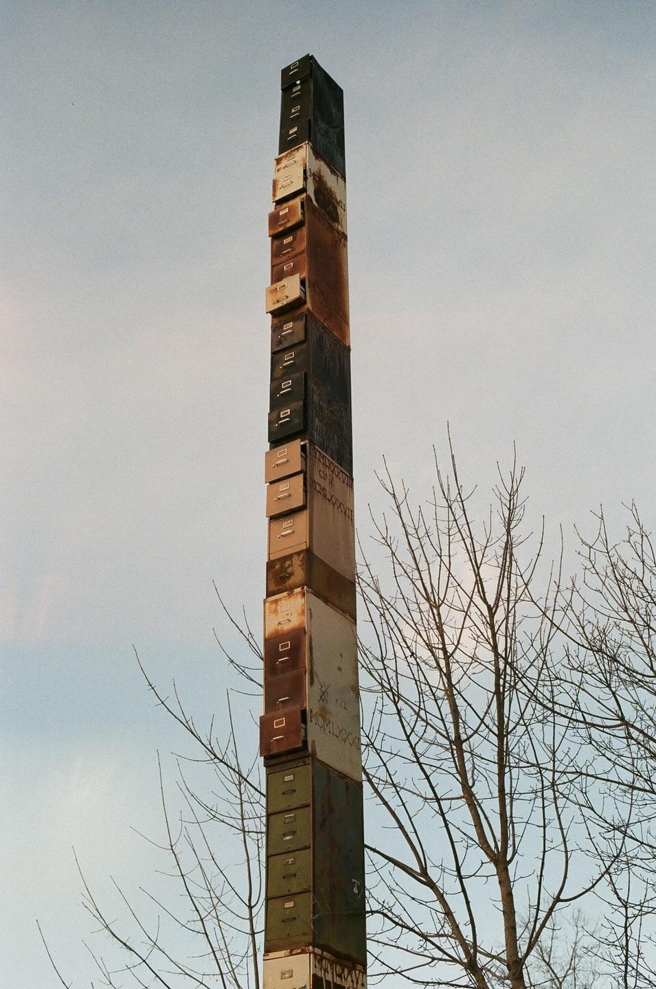 A tall sculpture made of stacked filing cabinets stands against a pale sky, with bare tree branches visible on the right.