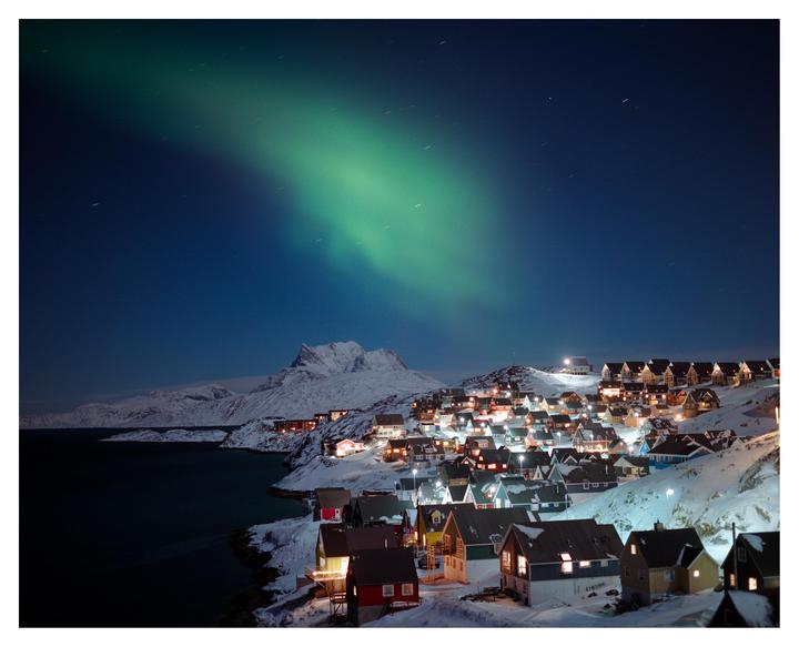 A green aurora borealis lights up the night sky over a snowy village on a coastline.