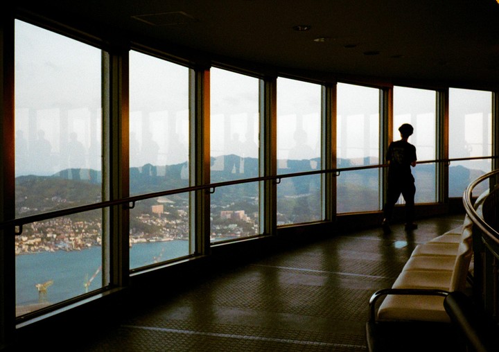 A person stands silhouetted against a large window overlooking a city and body of water.