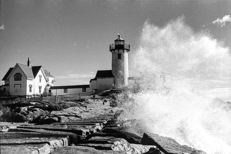 A large wave crashes against a rocky shore with a lighthouse and house in the background.