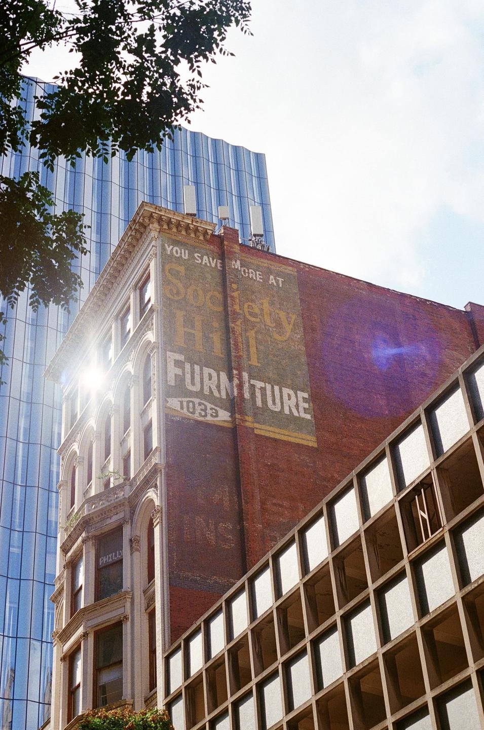 Sunlight shines on an old brick building with a painted advertisement for "Society Hill Furniture" next to a modern glass skyscraper.