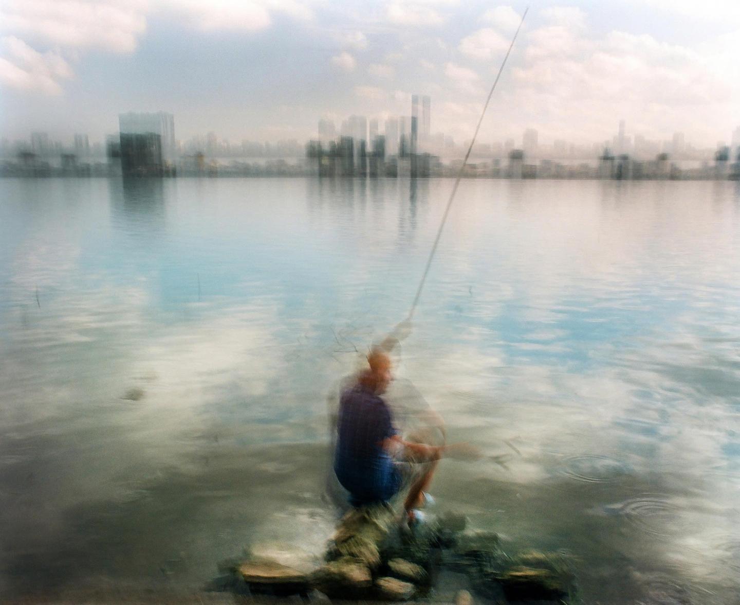 A man fishes in a body of water with a city skyline in the background.