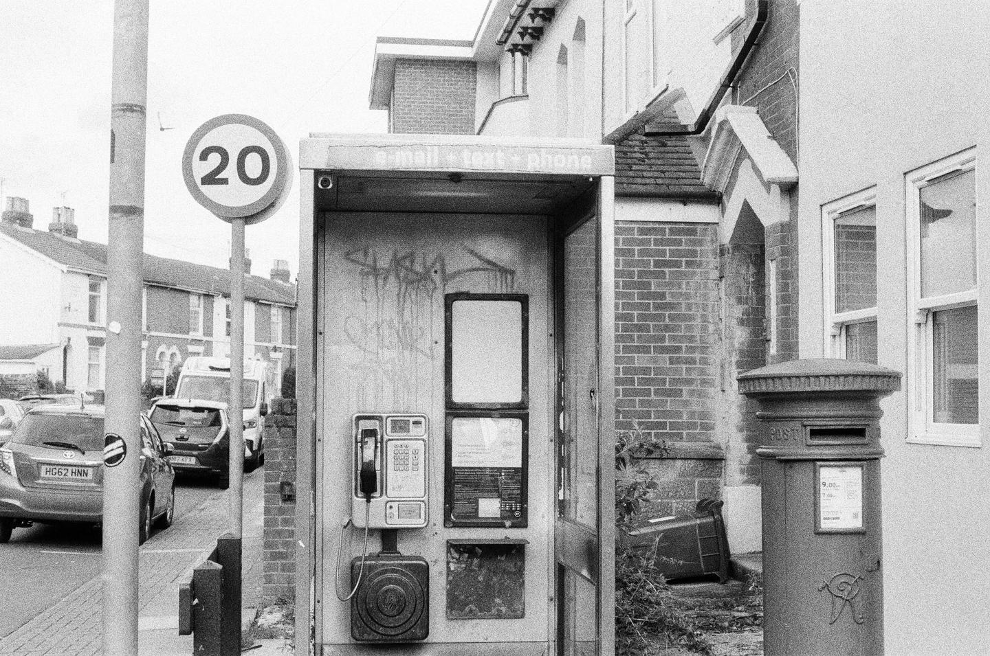 A black and white street scene shows a phone booth, a post box, and a speed limit sign.
