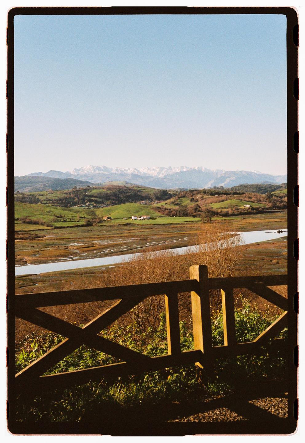 A wooden fence is in the foreground of a landscape with a river, green hills, and snow-capped mountains under a clear sky.